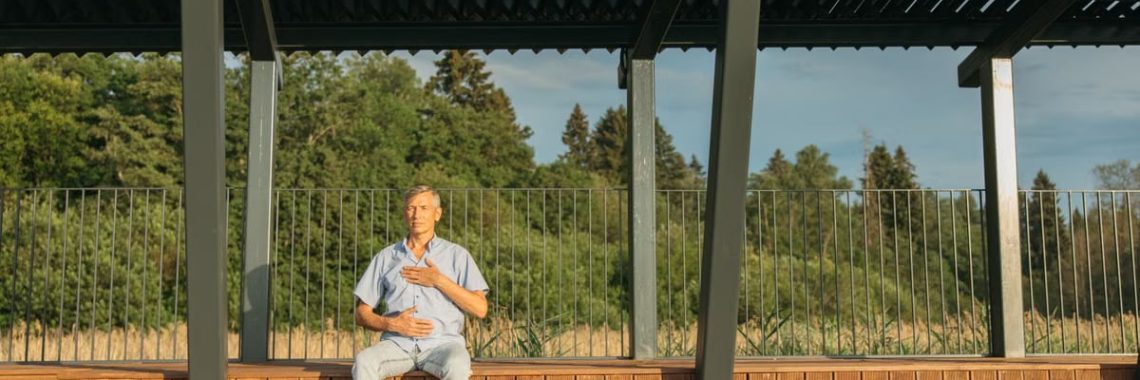 Middle-aged man practicing meditation outdoors on wooden bleachers under a metal structure in sunny weather.